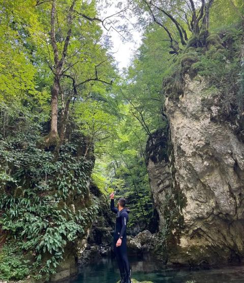 A canyoner in a black wetsuit standing at the edge of a clear turquoise pool in a wooded gorge, looking up at a pale limestone wall and green beech canopy.