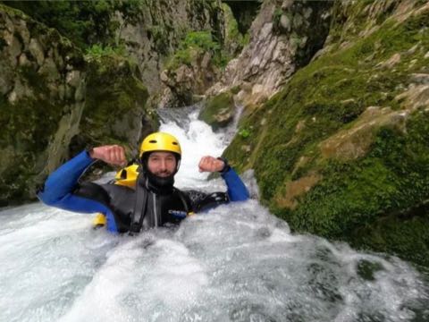 A canyoner in a blue and black wetsuit and yellow helmet sitting in the white water at the foot of a small cascade, arms raised, mossy boulders on either side.