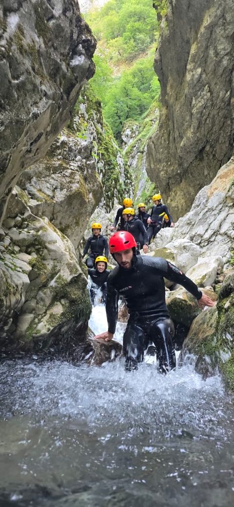 A line of six people in wetsuits and helmets stepping down a series of small cascades between tall grey walls, a guide in a red helmet leading.
