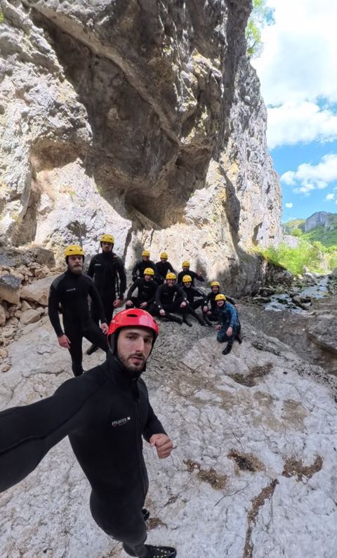 A group of about eleven people in wetsuits and yellow helmets resting on a broad pale limestone slab under an overhang, blue sky above, a guide in a red helmet in the foreground.