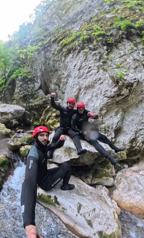 Three guides in red helmets and black wetsuits posing on a large pale boulder above a stream, with wet limestone and green vegetation behind them.