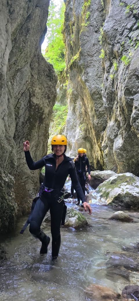 Two canyoners in black wetsuits and yellow helmets wading a shallow stretch of river between high grey limestone walls, one smiling towards the camera.
