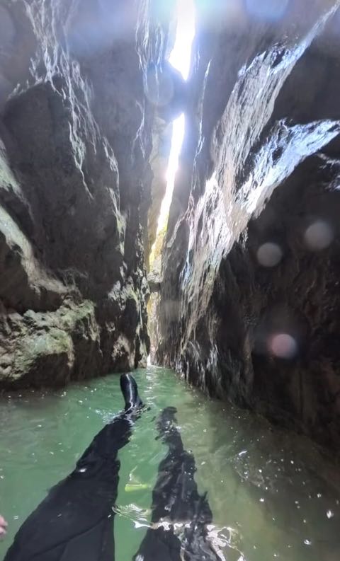 Swimming a narrow flooded slot in the canyon: dark wet walls rise close on both sides, green water ahead, and a thin bright line of sky far above.