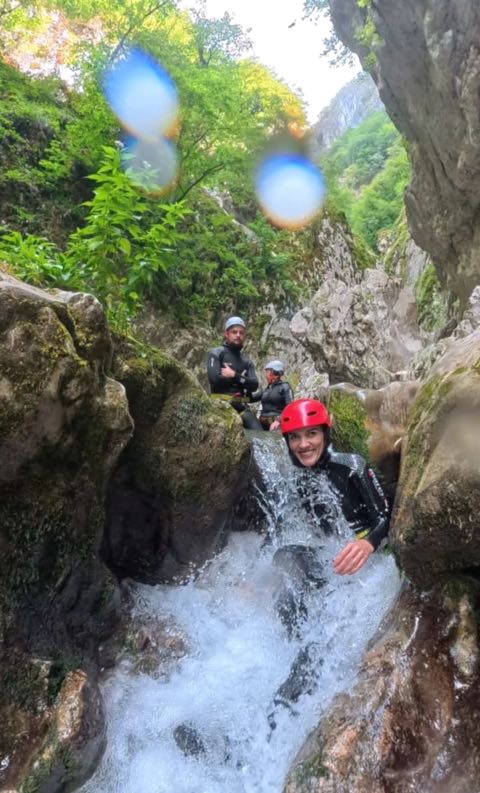 A woman in a red helmet coming down a short waterfall into the pool below, with two others in wetsuits watching from the rock above.