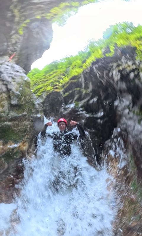 A canyoner in a red helmet coming down a white-water cascade between mossy walls, arms raised, spray filling the frame.
