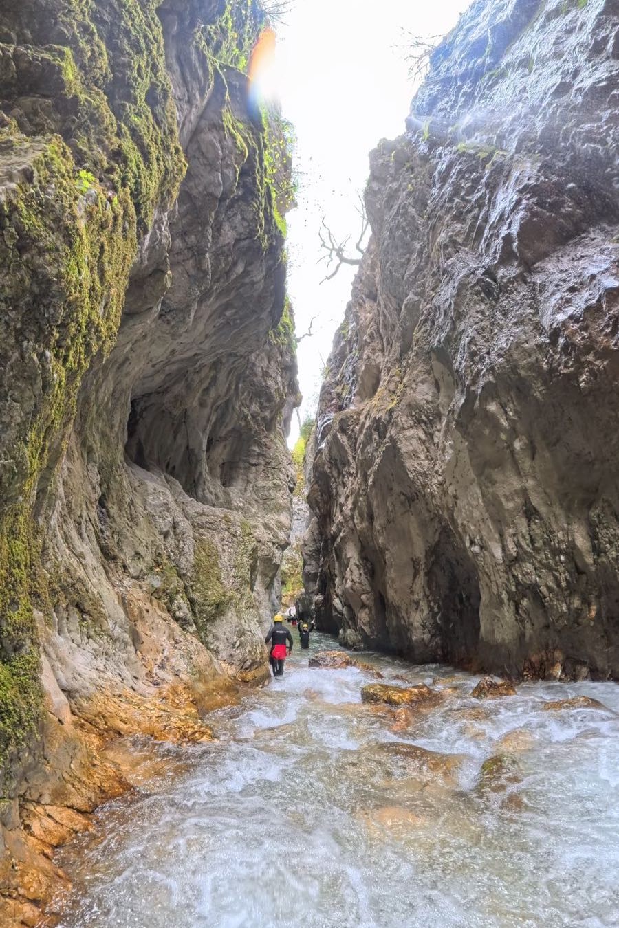 Looking up inside Nevidio canyon: sheer limestone walls on both sides almost meeting overhead, leaving a narrow strip of white sky. Three canyoners in helmets wade the pale green river far below.
