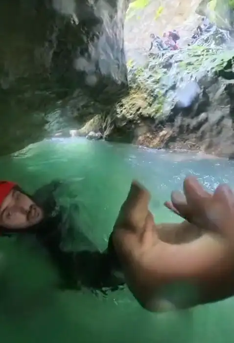 Point-of-view footage moving downstream through turquoise water towards a rock overhang.