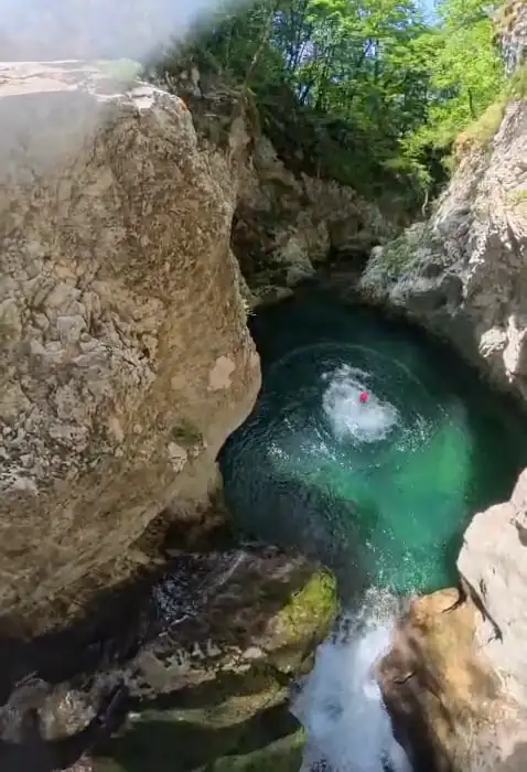 An emerald green pool beneath an enormous boulder, with a canyoner swimming across it.