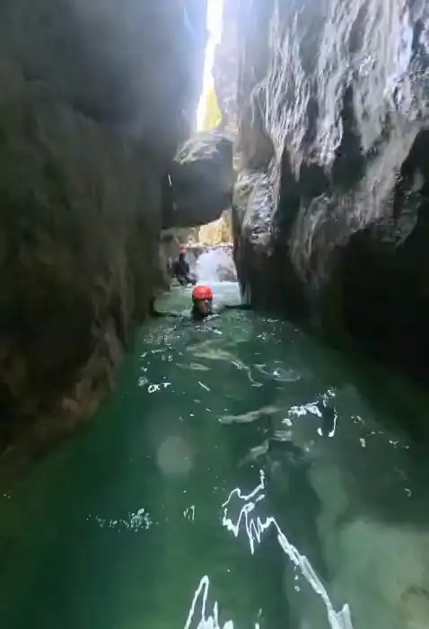 A canyoner in a red helmet swimming a narrow flooded slot, walls close on both sides and a bright seam of sky far above.